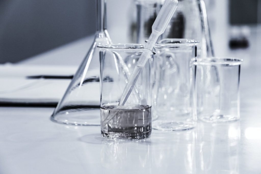 Transparent lab equipment on a white surface, including a beaker with liquid and a submerged pipette, alongside other empty glassware like a conical flask and beakers, suggesting a scientific or laboratory setting.