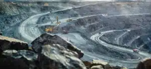 A large open-pit mine with dusty, terraced slopes and winding roads. Excavators and dump trucks operate on different levels. The atmosphere appears hazy, highlighting the scale and depth of the mining operation. Rocks are visible in the foreground.