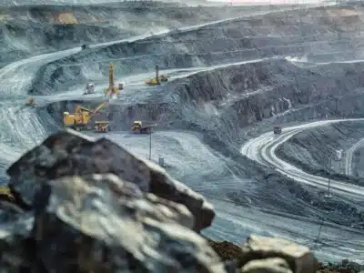 A large open-pit mine with dusty, terraced slopes and winding roads. Excavators and dump trucks operate on different levels. The atmosphere appears hazy, highlighting the scale and depth of the mining operation. Rocks are visible in the foreground.