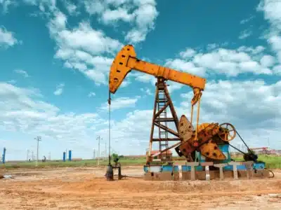 A large yellow oil pumpjack stands on barren land under a bright blue sky with scattered clouds. Power lines and industrial structures are visible in the background.