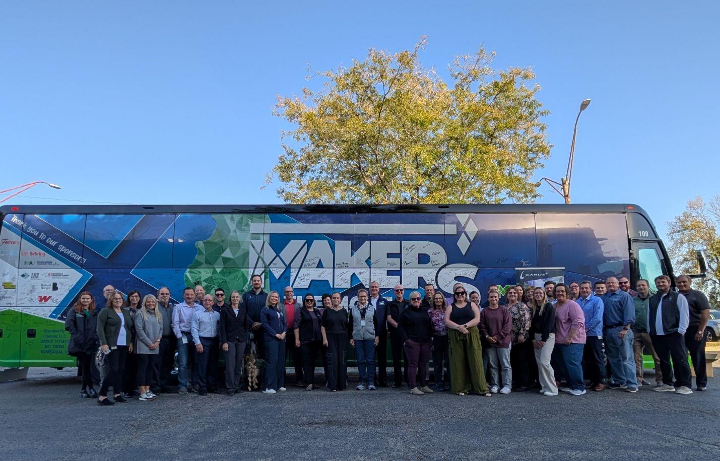 A large group of people stands in front of a colorful bus with 'Makers' written on its side. The bus is parked next to a tree under a clear blue sky. The group is diverse and appears to be posing for a group photo.