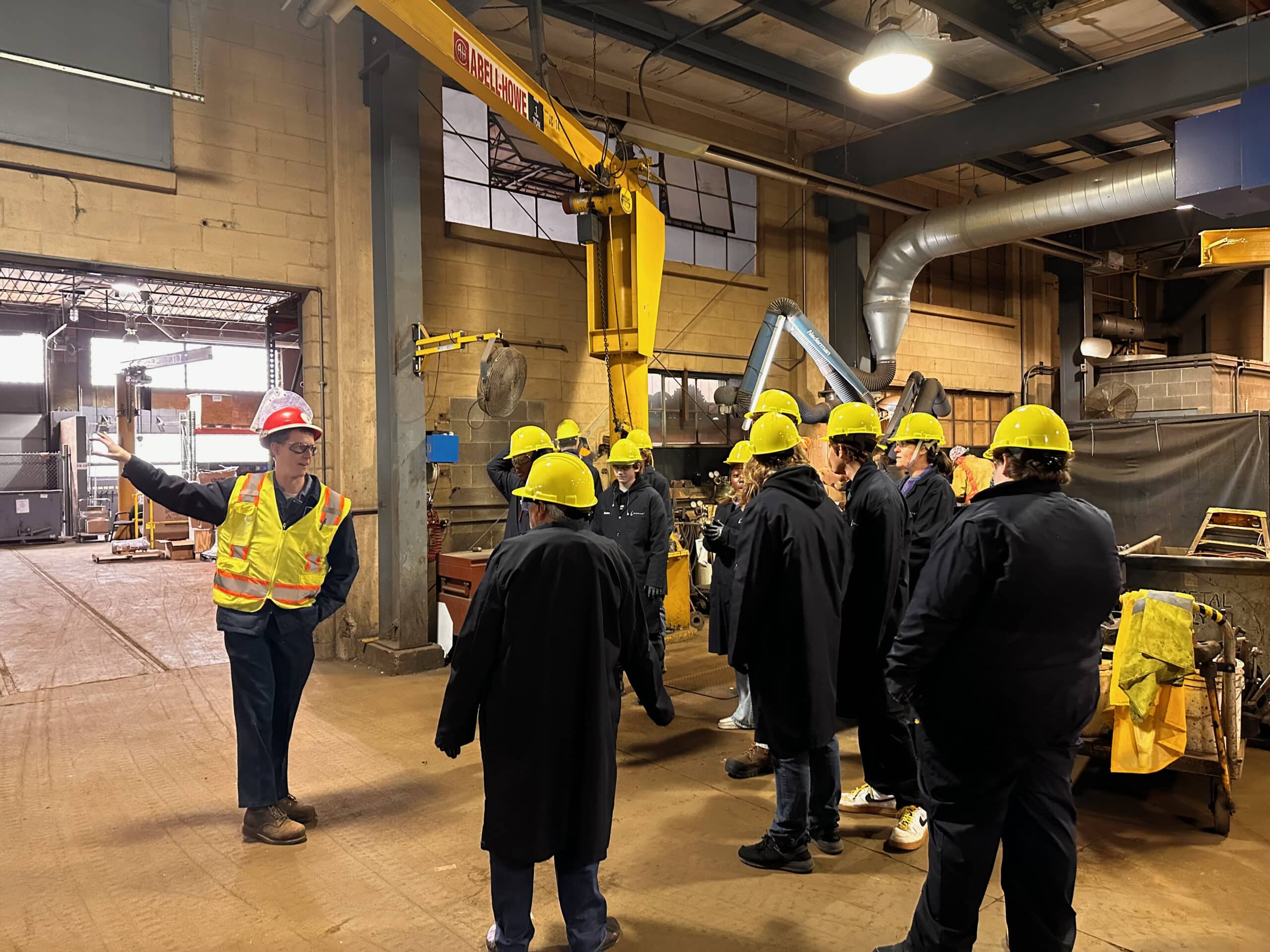 A group of people wearing yellow hard hats and dark coats stand inside a factory. A person in a red hard hat and neon safety vest gestures while talking to the group. Industrial equipment and large pipes are visible in the background.