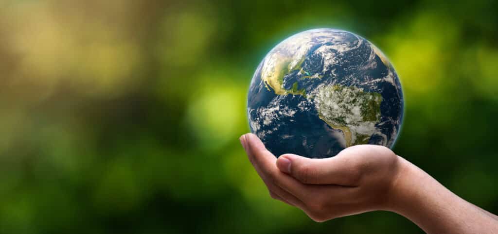 A hand gently holds a small, realistic model of Earth against a blurred green and yellow background, symbolizing environmental care and global responsibility. The continents and oceans are clearly visible in vibrant blue, green, and white.