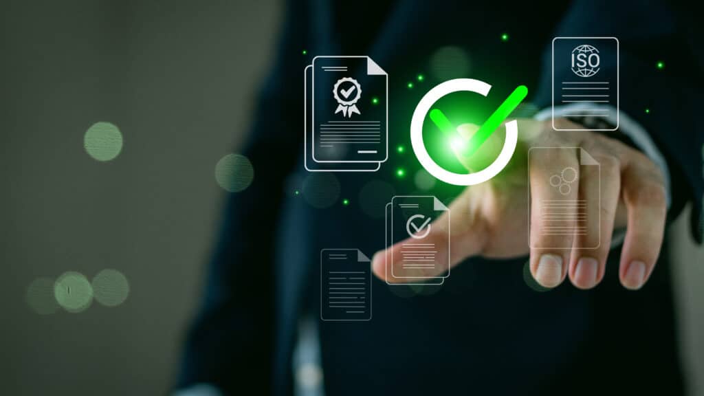 A person in a suit interacts with floating digital icons, including a large green check mark, a document with a check, and an ISO certification symbol. The background is blurred, emphasizing the focus on the hand and icons.