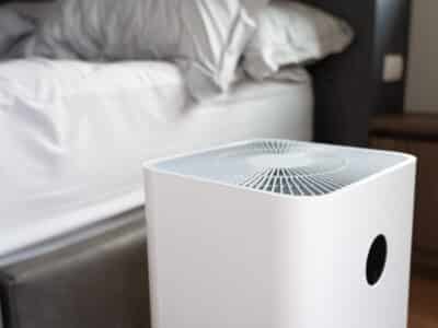 A white, modern air purifier is placed in a bedroom next to a bed with white linens. The device features a top vent and a circular sensor on the front. The room has a cozy, minimalistic decor.