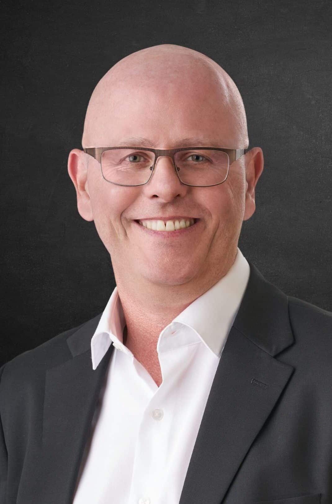 A man wearing glasses smiles at the camera. He is dressed in a black suit jacket over a white collared shirt. The background is a solid dark color.