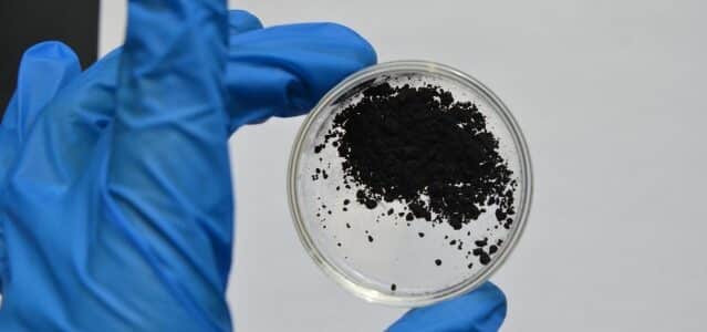 A gloved hand holds a small Petri dish containing a black powdery substance against a neutral background.