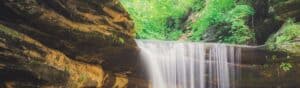 A serene waterfall cascades over smooth rocks, surrounded by lush green foliage. Sunlight filters through the trees, illuminating the water and creating a tranquil, natural scene.