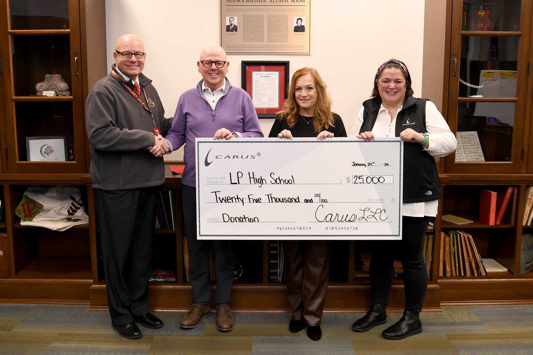 Four people stand indoors holding a large ceremonial check for $25,000 made out to LP High School. They are positioned in front of a wooden bookshelf with framed items, smiling for the photo.