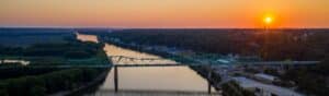 Aerial view of a green truss bridge spanning a wide river during sunset. The sun is low on the horizon, casting warm colors over the water. Lush green trees line the riverbanks, and buildings are visible in the distance.