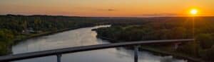 A wide river flows beneath a bridge at sunset, surrounded by lush green forests. The sun sets on the horizon, casting a warm glow over the landscape. A cluster of buildings and trees is visible along the riverbank.