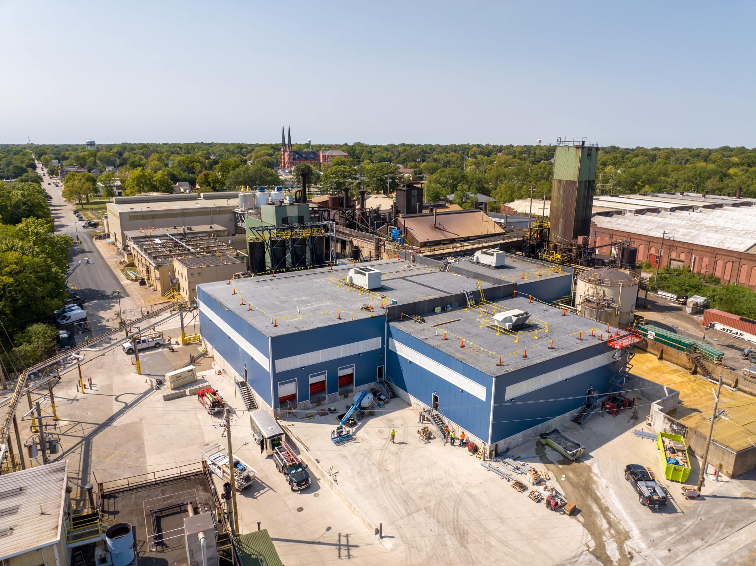 Aerial view of an industrial complex with large flat-roofed buildings and surrounding equipment. The main building is blue and gray. Several vehicles and construction materials are scattered around the premises. Trees and a town are visible in the distance.