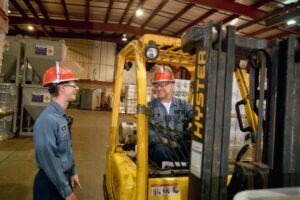 Two workers wearing red hard hats and blue uniforms are in a warehouse. One operates a yellow forklift labeled "HYSTER," while the other stands nearby, both smiling and engaging in conversation. Stacks of goods and equipment surround them.