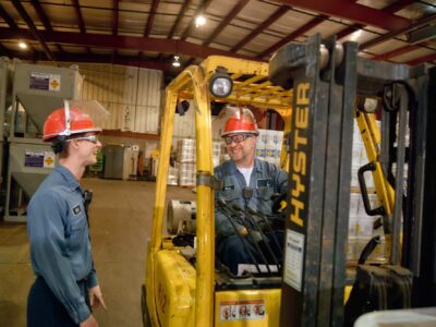 Two workers wearing red hard hats and blue uniforms are in a warehouse. One operates a yellow forklift labeled "HYSTER," while the other stands nearby, both smiling and engaging in conversation. Stacks of goods and equipment surround them.