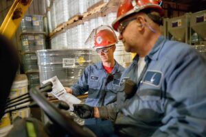 Two workers wearing red hard hats and blue uniforms are in a warehouse filled with stacked barrels. One is seated, holding a pamphlet, while the other leans over, both focused on the document. Safety gear and industrial equipment surround them.