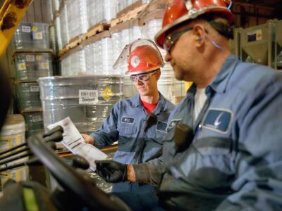 Two workers wearing red hard hats and blue uniforms are in a warehouse filled with stacked barrels. One is seated, holding a pamphlet, while the other leans over, both focused on the document. Safety gear and industrial equipment surround them.