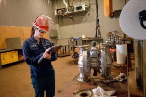 A person wearing safety gear and glasses stands in an industrial workshop, writing on a clipboard. They are next to a large metal pump on a workbench. Various tools and equipment are visible in the background.