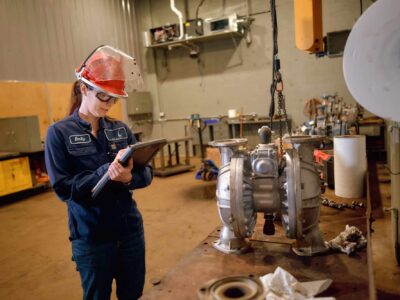 A person wearing safety gear and glasses stands in an industrial workshop, writing on a clipboard. They are next to a large metal pump on a workbench. Various tools and equipment are visible in the background.