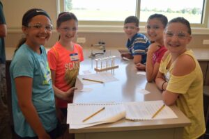 Five children wearing safety goggles smile while gathered around a lab table with papers and pencils. A test tube rack is in the background, and they appear to be in a science classroom with a window showing a green landscape outside.