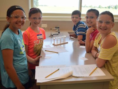 Five children wearing safety goggles smile while gathered around a lab table with papers and pencils. A test tube rack is in the background, and they appear to be in a science classroom with a window showing a green landscape outside.