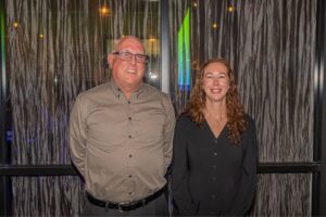 A man and a woman are standing side by side, smiling. They are in front of a textured wall with decorative lights. The man is wearing glasses and a gray shirt, while the woman has long curly hair and is wearing a black blouse.