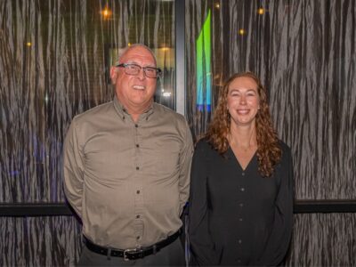 A man and a woman are standing side by side, smiling. They are in front of a textured wall with decorative lights. The man is wearing glasses and a gray shirt, while the woman has long curly hair and is wearing a black blouse.