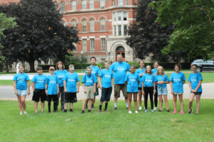 A group of children and adults wearing matching blue shirts stand on a grass field, posing for a photo. A red brick building is in the background, along with trees and a parked car.