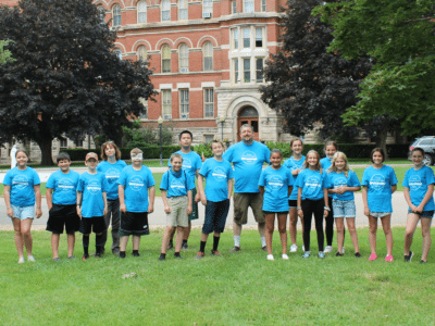 A group of children and adults wearing matching blue shirts stand on a grass field, posing for a photo. A red brick building is in the background, along with trees and a parked car.