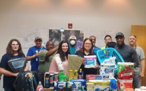 A group of people smiling and posing with cleaning supplies and toiletries, including paper towels, tissues, and detergent. There are maps on the wall behind them and assorted items on the table in front.