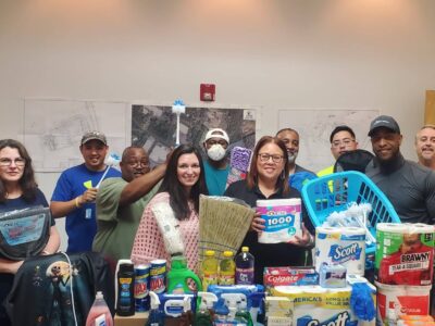 A group of people smiling and posing with cleaning supplies and toiletries, including paper towels, tissues, and detergent. There are maps on the wall behind them and assorted items on the table in front.