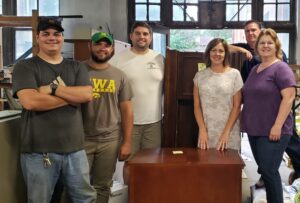 A group of six people stand smiling indoors around a wooden cabinet. They are in a room with large windows and various items in the background. Four are standing and two are partially leaning on the furniture.