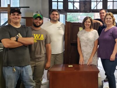 A group of six people stand smiling indoors around a wooden cabinet. They are in a room with large windows and various items in the background. Four are standing and two are partially leaning on the furniture.