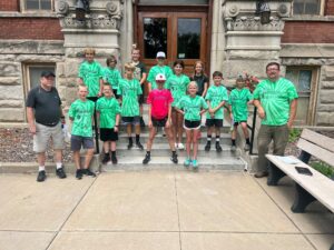 A group of children and two adults stand on the steps of a brick building. The children wear matching green tie-dye shirts, and one child in the center wears a red shirt. The adults are dressed casually. The building has ornate stonework and wooden doors.