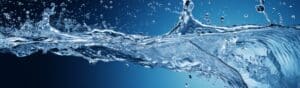 Close-up of water splashing against a dark blue background, showing droplets in mid-air. The water forms dynamic shapes and patterns, highlighting its clarity and motion.