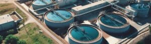 Aerial view of a wastewater treatment plant featuring several round tanks filled with blue water. The facility has interconnected pipes and is surrounded by pathways and green foliage. Buildings are visible in the background.