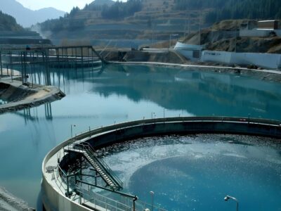 A serene water treatment facility nestled in a mountainous landscape. Circular basins filled with clear blue water reflect the surrounding hills and sky. Various structures and pathways border the basins, with trees lining the distant background.