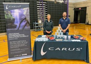 A man and woman stand behind a table with a Carus banner and branded materials. The table has cups, candy, pens, and informational pamphlets. The setting appears to be a gymnasium with bleachers in the background.