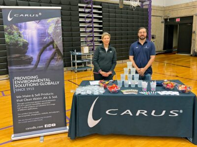 A man and woman stand behind a table with a Carus banner and branded materials. The table has cups, candy, pens, and informational pamphlets. The setting appears to be a gymnasium with bleachers in the background.