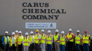 A group of people wearing hard hats and high-visibility vests stand in front of a building with the sign "Carus Chemical Company." A smaller banner reads "...working with Responsible Care.