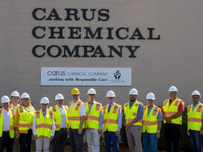 A group of people wearing hard hats and high-visibility vests stand in front of a building with the sign "Carus Chemical Company." A smaller banner reads "...working with Responsible Care.