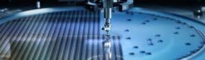 Close-up view of a semiconductor manufacturing process. A robotic arm operates over a large silicon wafer, placing tiny components with precision. The wafer has numerous reflective microchips arranged in a grid pattern.