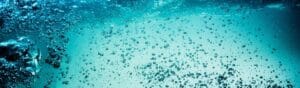 Underwater scene with numerous air bubbles rising through clear, turquoise water. Light filters from above, creating a bright, refreshing ambiance. Some larger bubbles are clustered in the bottom left corner.