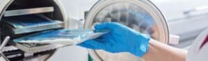 A gloved hand placing a sealed, sterile package into an open autoclave machine with a circular door, used for sterilizing equipment. The autoclave's interior is visible, showing a metal rack.