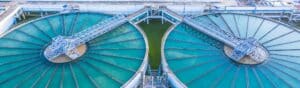 Aerial view of two large circular water treatment tanks with aquamarine-colored water. Metal walkways extend across both tanks, which are part of an industrial facility. Structures and pathways are visible in the background.