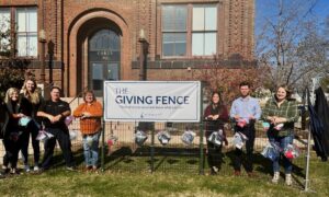 A group of people stands in front of a building beside a fence adorned with bags. A banner on the fence reads "The Giving Fence" with a tagline about taking what you need. The people are smiling and standing on grass with trees nearby.