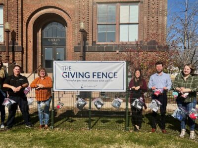 A group of people stands in front of a building beside a fence adorned with bags. A banner on the fence reads "The Giving Fence" with a tagline about taking what you need. The people are smiling and standing on grass with trees nearby.