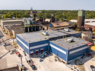 Aerial view of an industrial building with a flat roof, surrounded by machinery and other structures. Workers and vehicles are present around the site. Green trees and a church with spires are visible in the background under a clear sky.