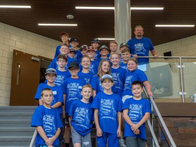 A group of children and two adults are posing for a photo on indoor stairs. They are all wearing matching blue t-shirts with white text. The setting appears to be inside a modern building with a wooden ceiling and glass railings.