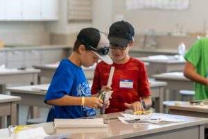 Two young boys in a classroom work together on a project. One wears a blue shirt and cap, while the other is in a red shirt and black cap. They are focused on assembling an object using small materials spread on their table.