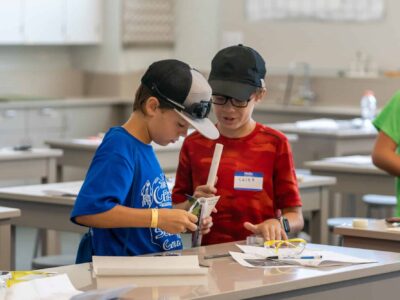 Two young boys in a classroom work together on a project. One wears a blue shirt and cap, while the other is in a red shirt and black cap. They are focused on assembling an object using small materials spread on their table.